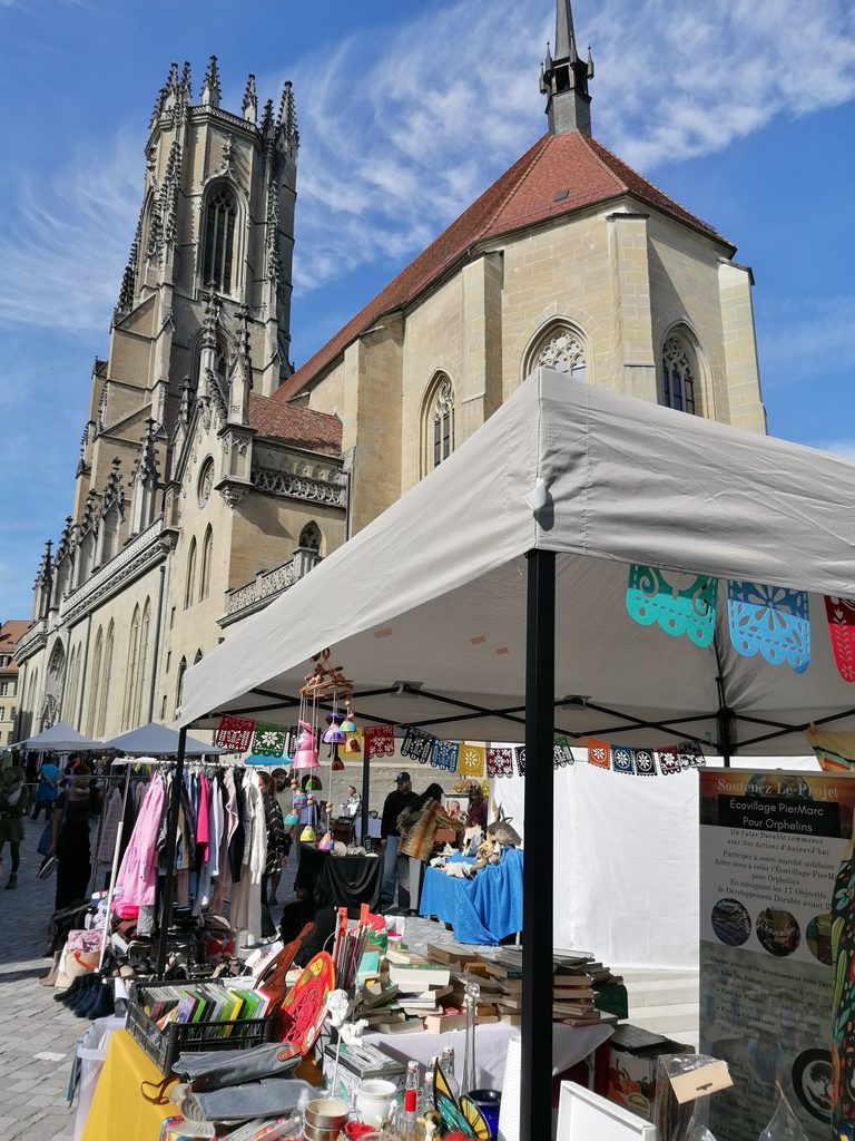Stand de l’ONG PierMarc Suisse au Marché aux Puces de Fribourg, proche de la cathédrale, avec livres et objets proposés aux visiteurs.