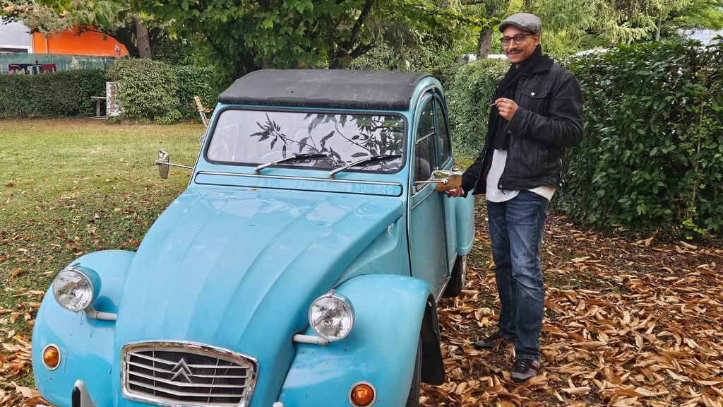 Babacar, bénévole de l’ONG PierMarc Suisse, à côté d’une Citroën 2CV bleue, sur un tapis de feuilles mortes à Meyrin.